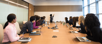 Room-full-of-professionals-round-a-conference-table-with-woman-writing-on-a-whiteboard