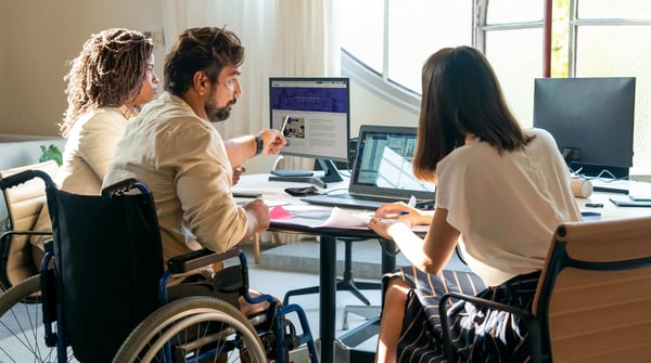 Two women and a man in a wheelchair looking at a laptop and a monitor. 